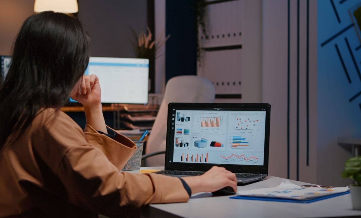 Woman working at a desk reviewing charts and graphs on a laptop screen in an office environment.