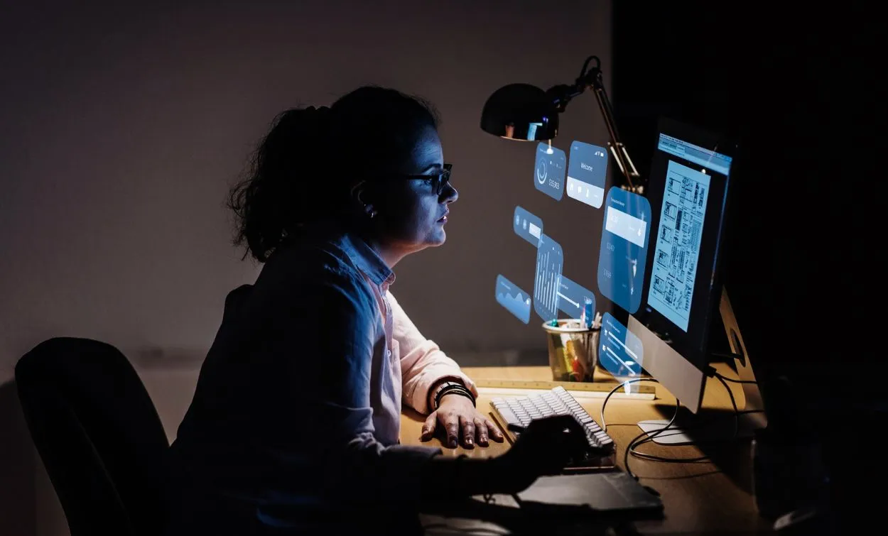 Woman learning software development and working on a computer at a desk in a dark workspace.